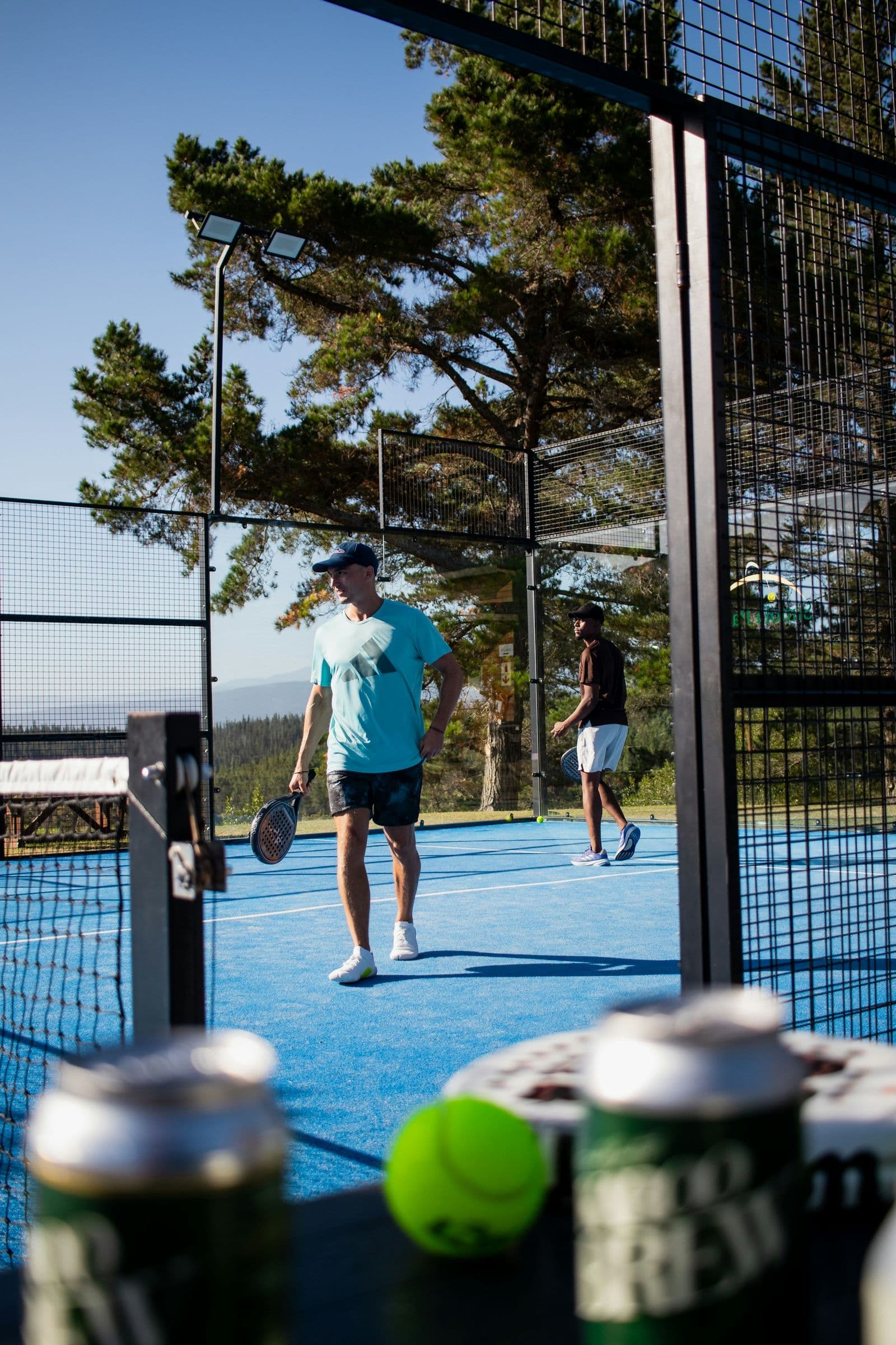 Two players on an outdoor padel court, with drinks and a ball in the foreground