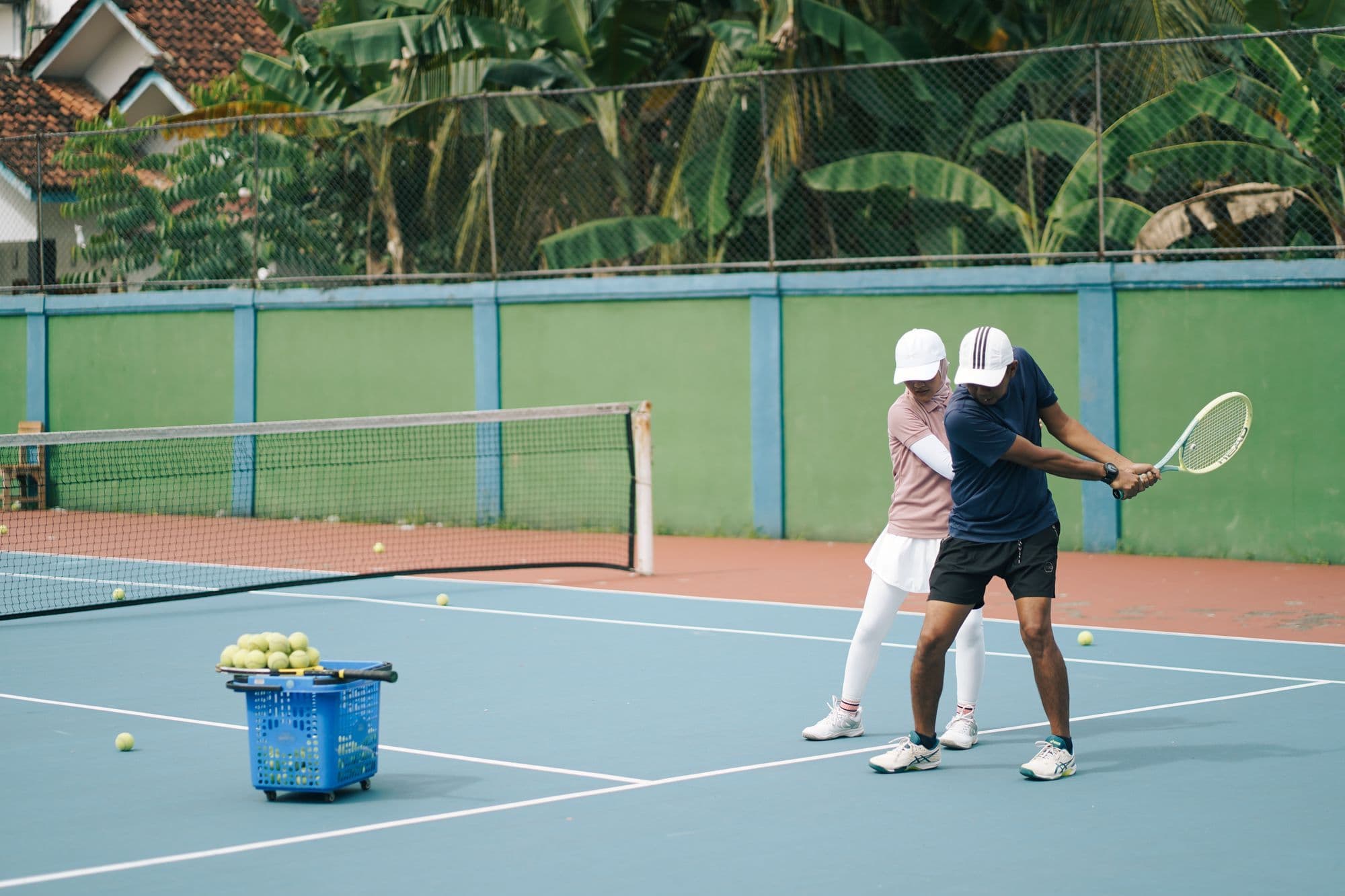 A tennis coach guiding a player's forehand on an outdoor hard court