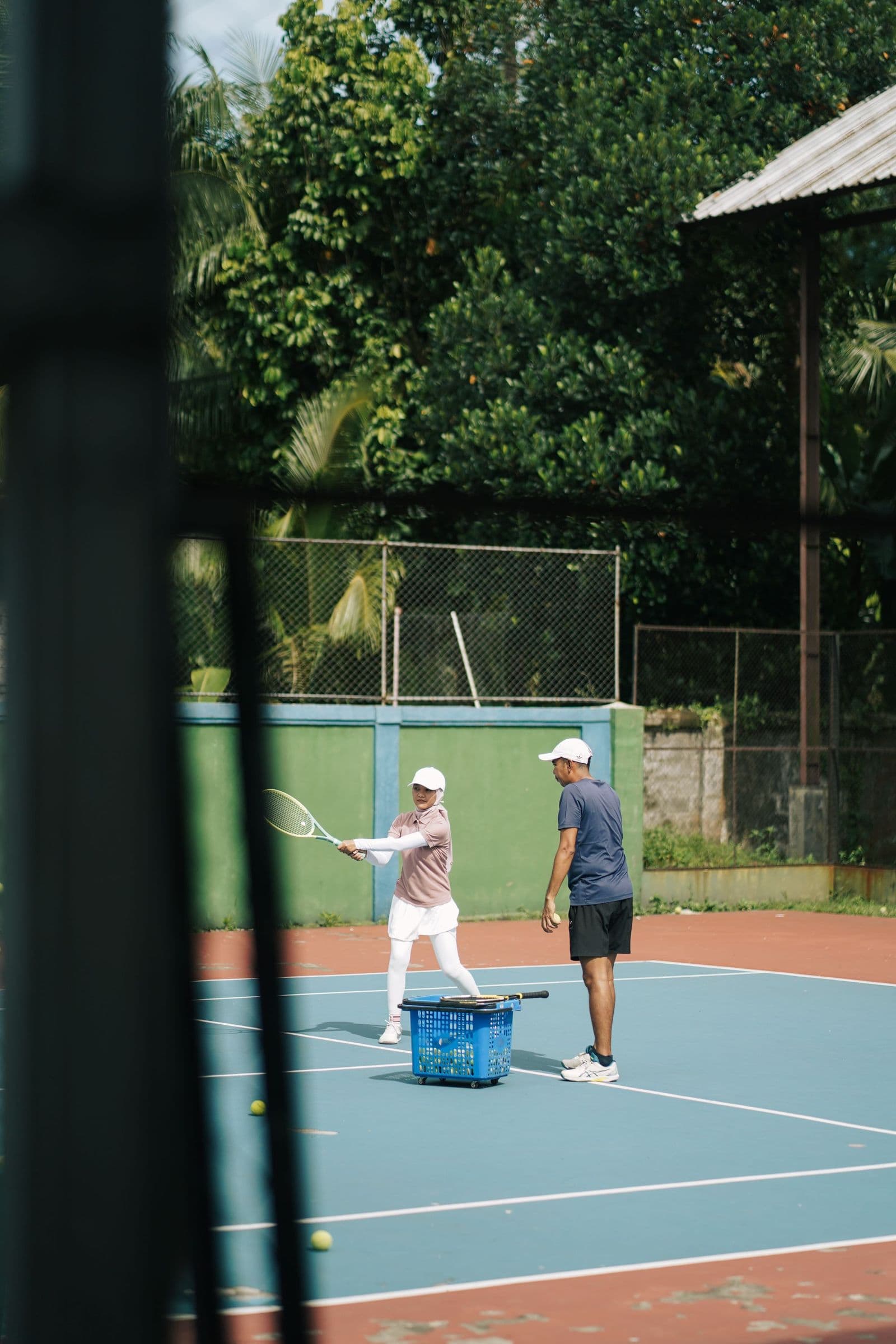 A coach and player during a private tennis lesson, seen through the court fence