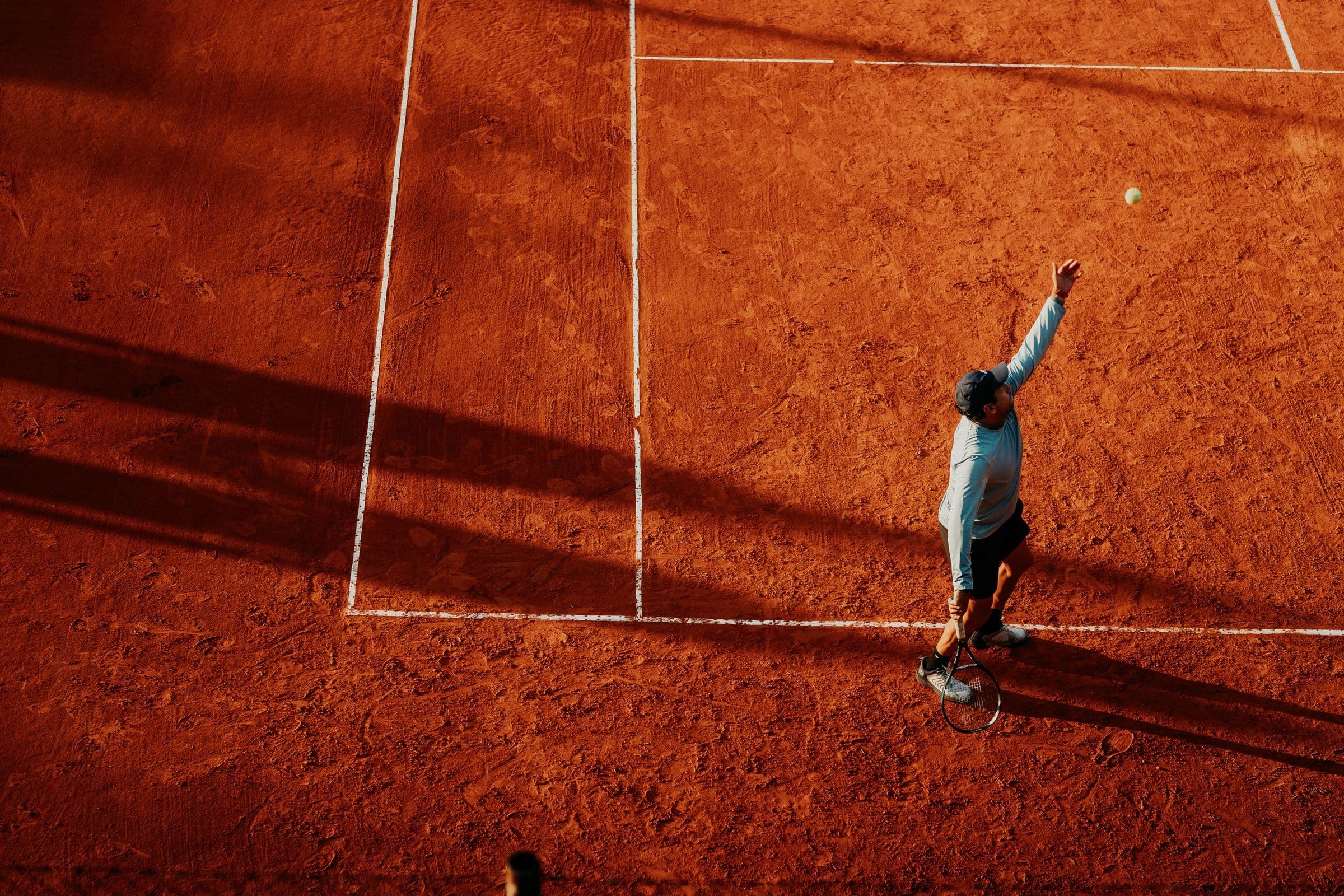A player serving on a sunlit clay court, seen from above