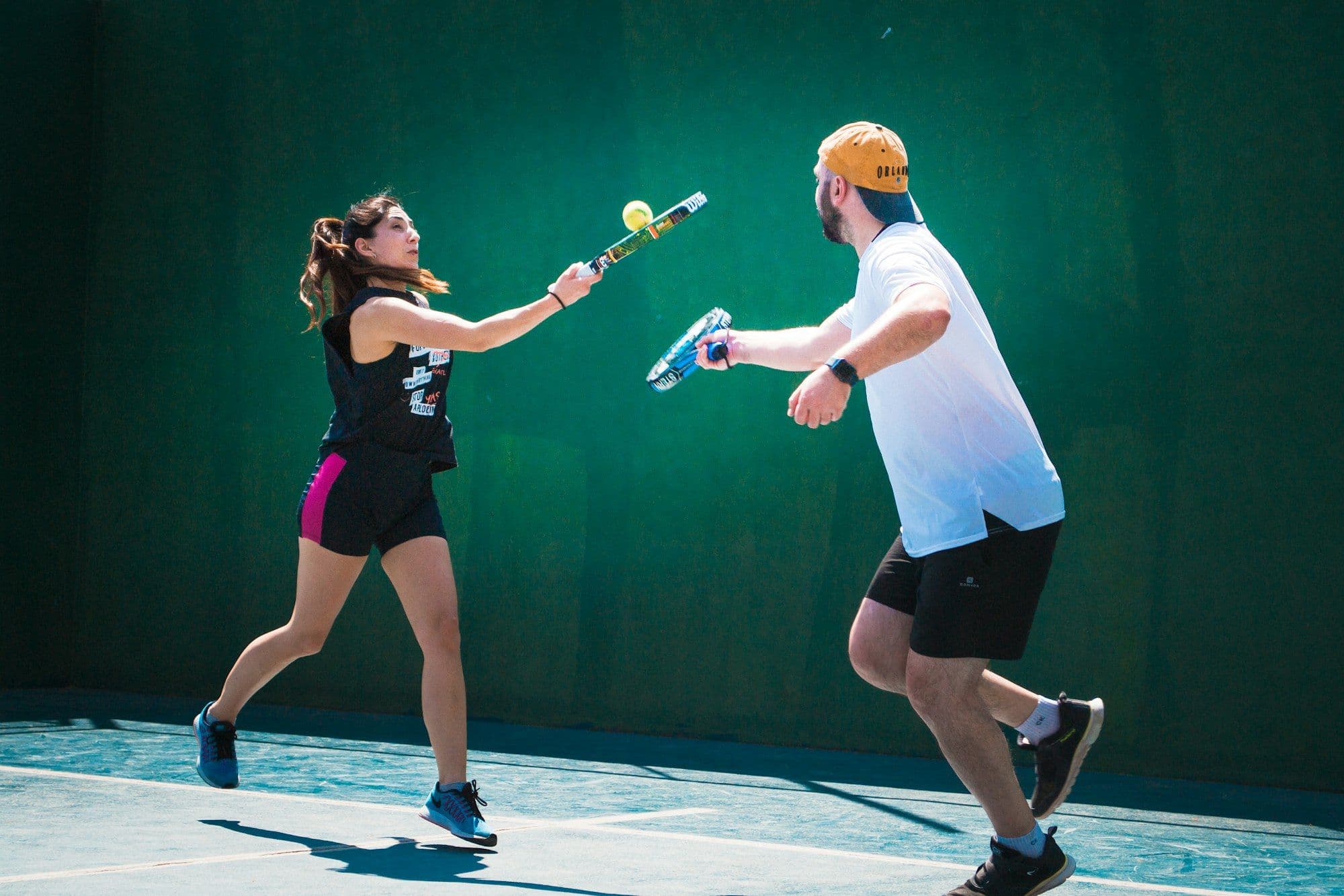 A padel coach feeding a volley to a player against a green court wall