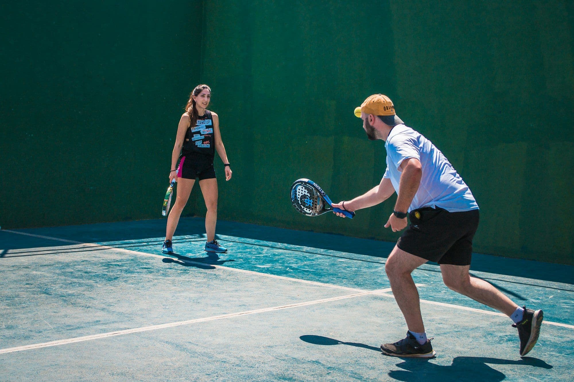 A padel coach and player rallying in front of a green wall
