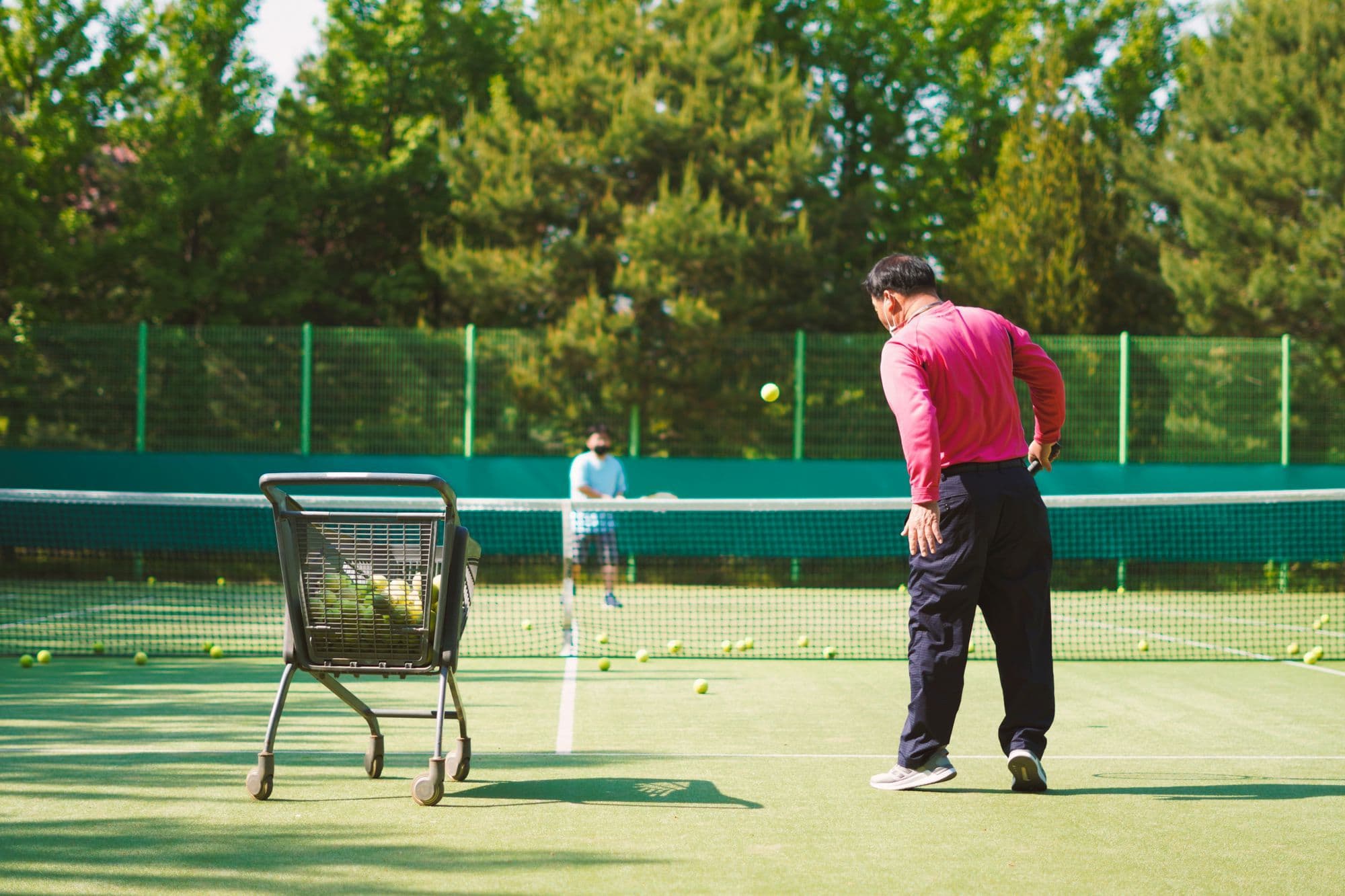 A tennis coach with a ball cart on a green court, trees behind